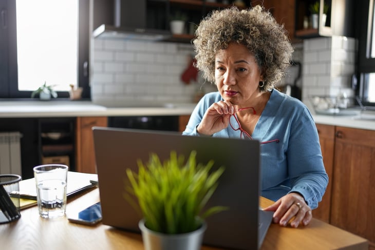 A concerned woman with curly gray hair reviews paperwork at her kitchen table, surrounded by a laptop, calculator, and papers